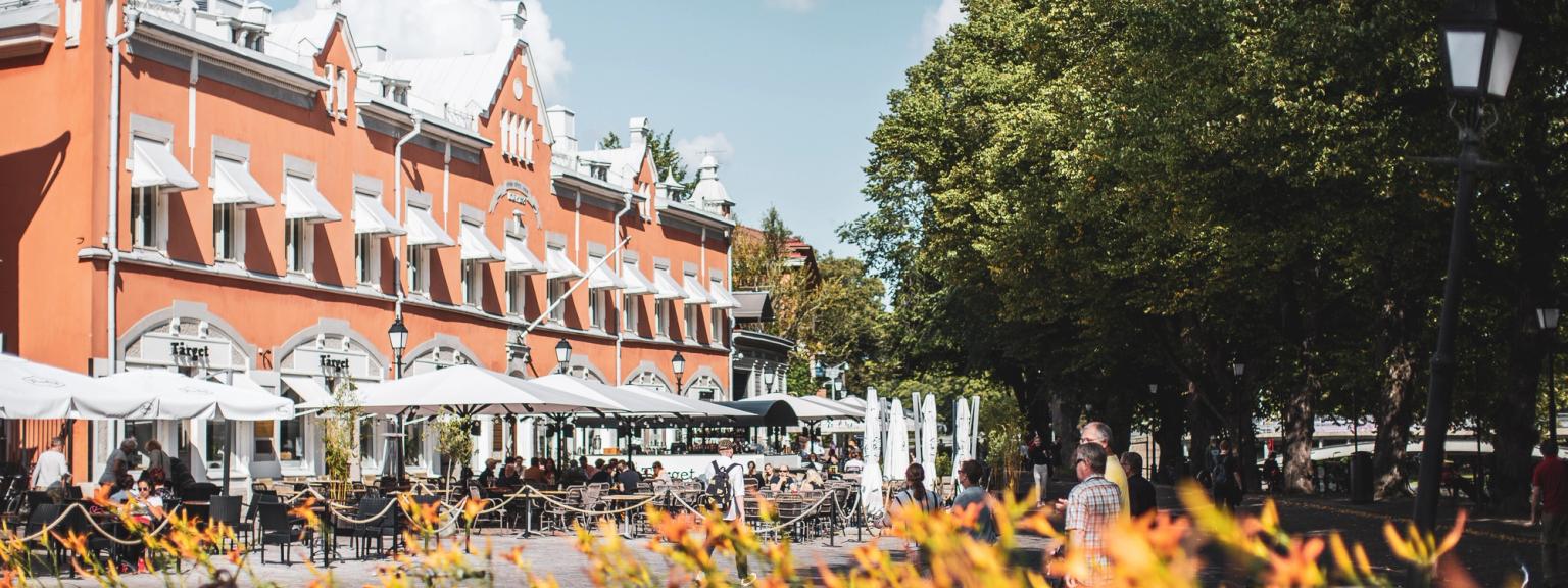 Ulkokahviloita, joissa ihmiset ruokailevat oranssin rakennuksen edessä, valkoisten markiisien alla, sinisen taivaan alla, puiden ja oranssien kukkien keskellä.