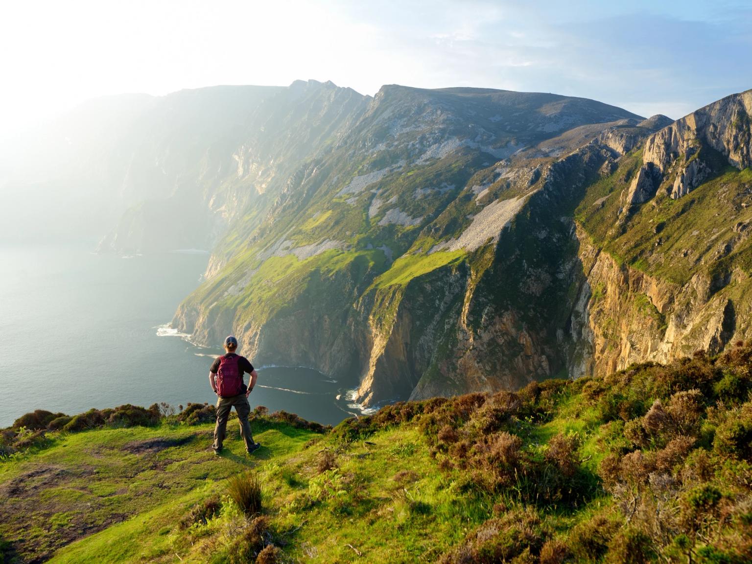 Person on a grassy cliff overlooking a vast ocean and dramatic sea cliffs.