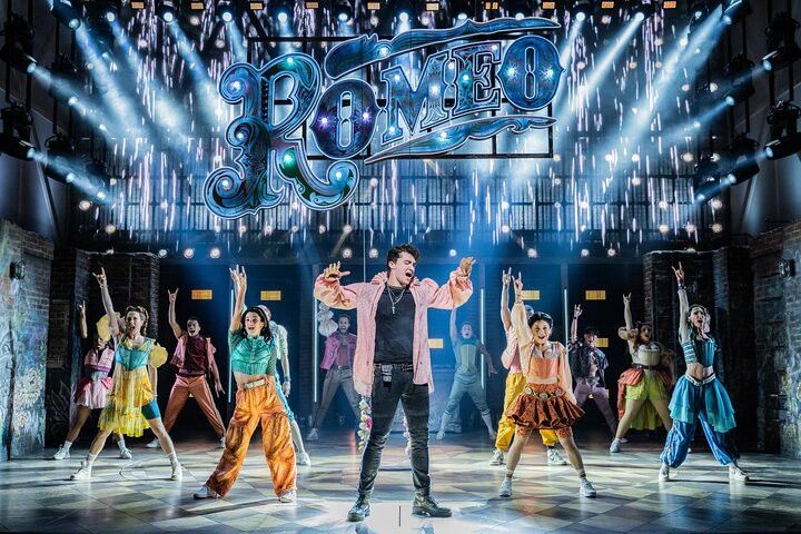 Die Besetzung des Musicals „Romeo“ tritt auf einer hell beleuchteten Bühne mit funkelnden Effekten und einem großen Schild mit der Aufschrift „ROMEO“ auf.