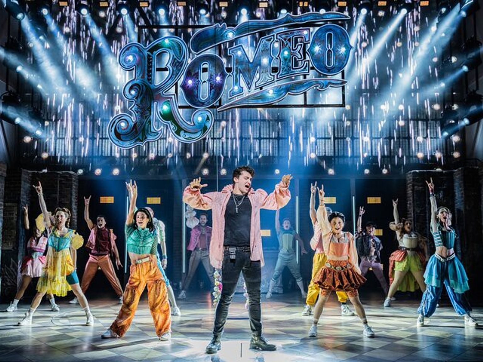 Die Besetzung des Musicals „Romeo“ tritt auf einer hell beleuchteten Bühne mit funkelnden Effekten und einem großen Schild mit der Aufschrift „ROMEO“ auf.