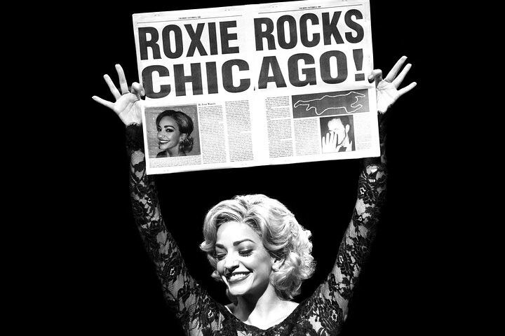 Schwarz-weiß-Bild einer lächelnden Frau mit blonden Locken, die eine Zeitung über ihrem Kopf hält, auf der „ROXIE ROCKS CHICAGO!“ steht.