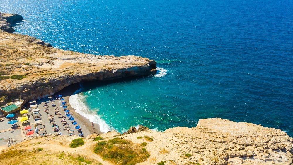 Luchtfoto van een afgelegen strand met turkoois water en kleurrijke parasols, omgeven door rotsachtige kliffen. Rethymnon, Griekenland
