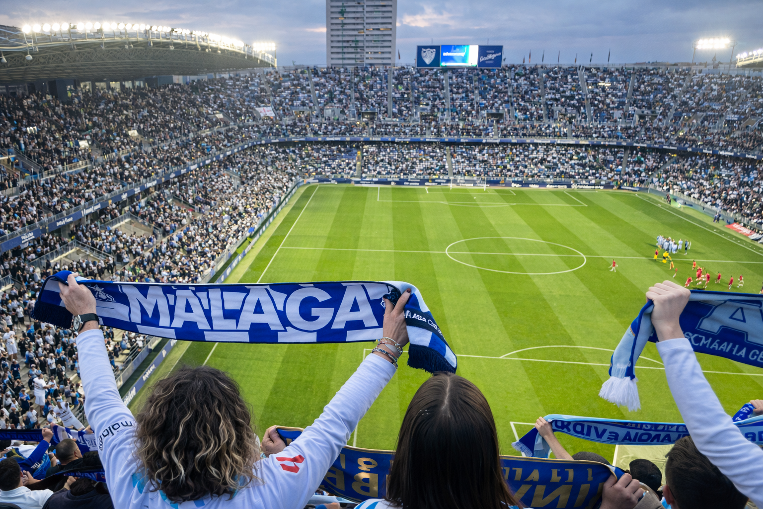 Vista desde detrás de los aficionados que sostienen bufandas de "Málaga" en un estadio de fútbol lleno.
