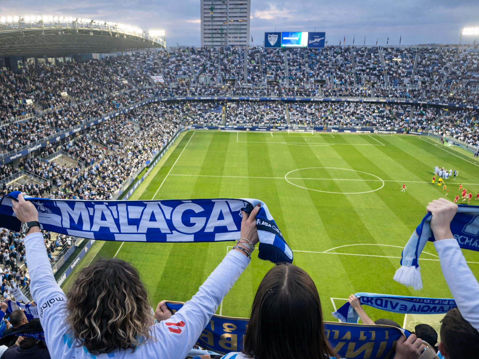 View from behind fans holding "Malaga" scarves in a packed soccer stadium.