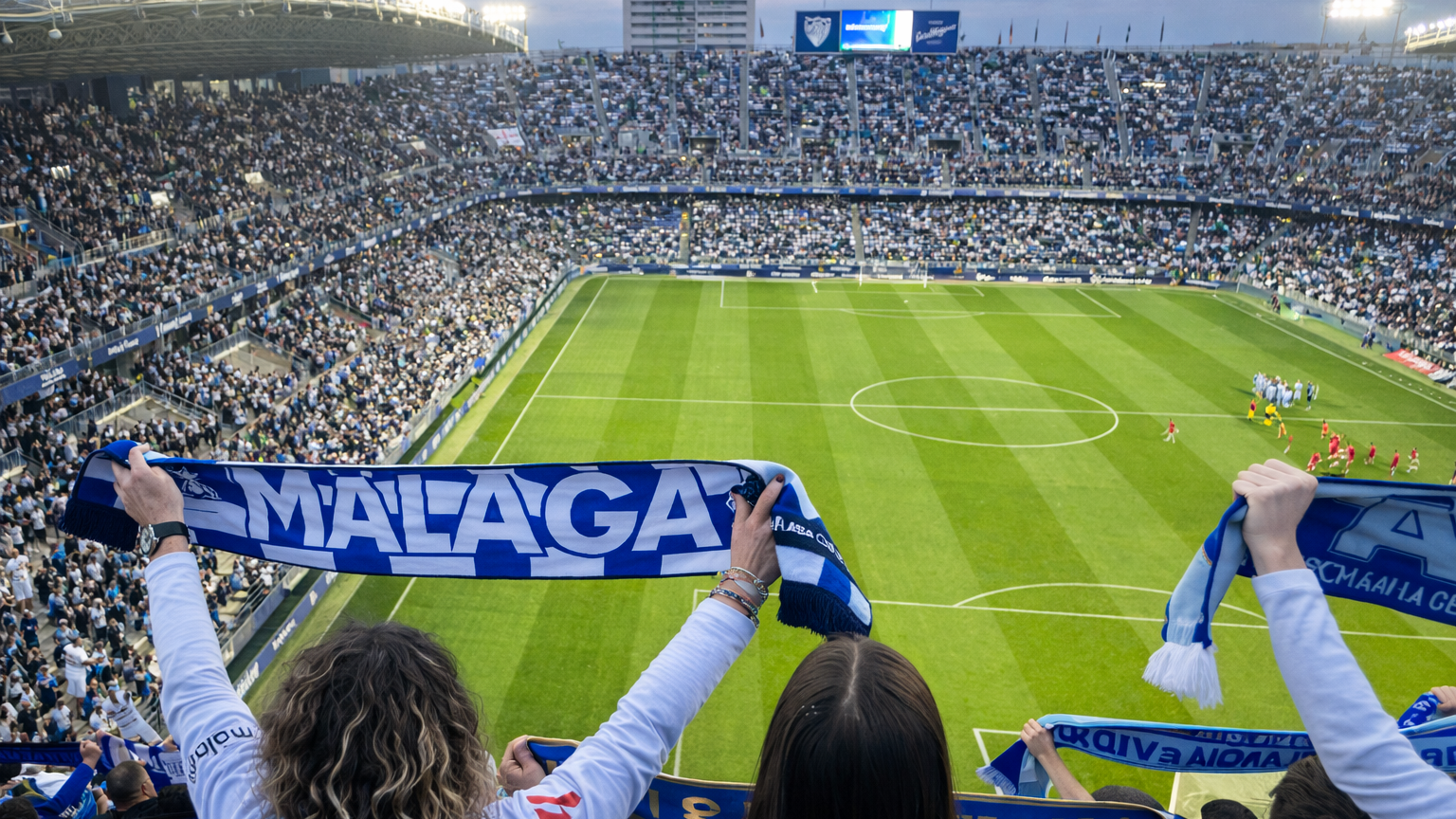 Vista desde detrás de los aficionados que sostienen bufandas de "Málaga" en un estadio de fútbol lleno.