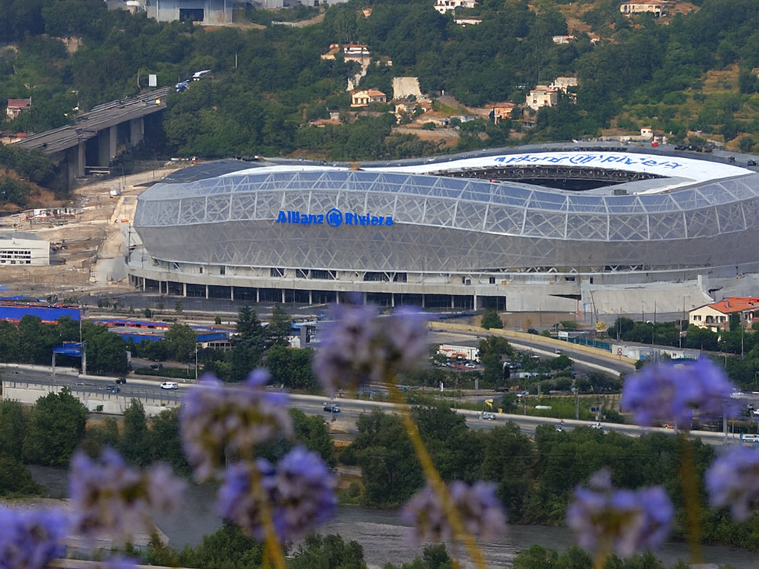Estádio Allianz Riviera cercado por colinas arborizadas com casas e estradas, visto através de flores roxas desfocadas em primeiro plano.
