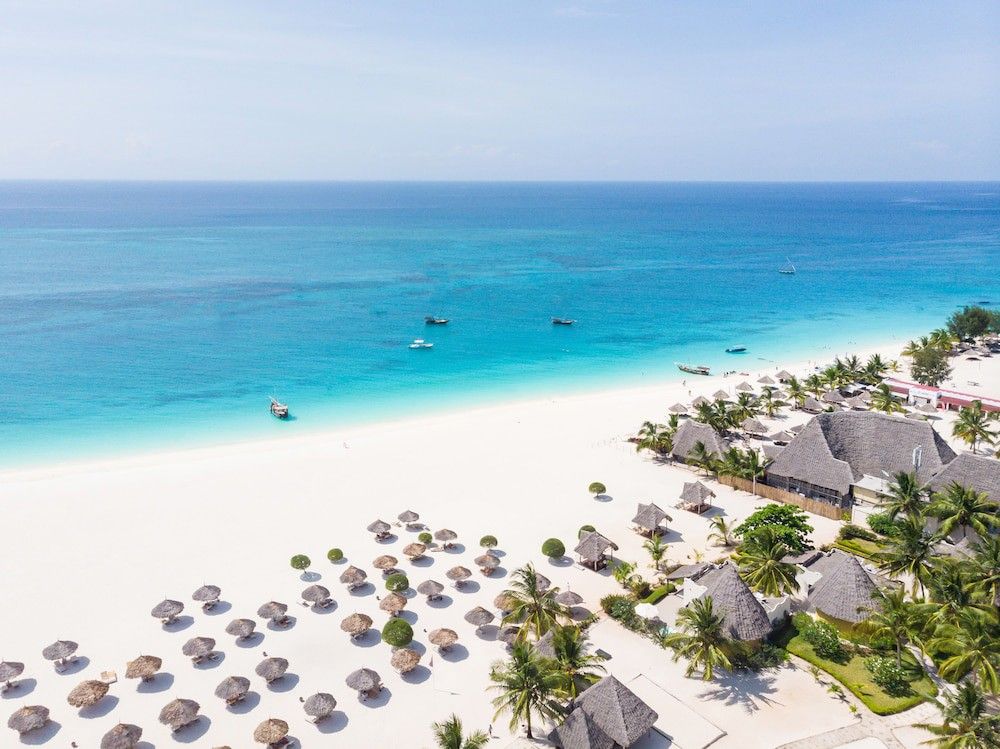 Aerial view of a white sand beach with turquoise water, boats, thatched umbrellas, and a resort.