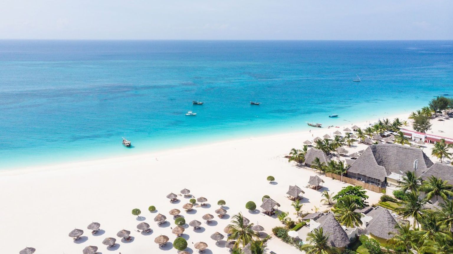 Flygfoto över en vit sandstrand med turkost vatten, båtar, halmtakparaplyer och en resort.