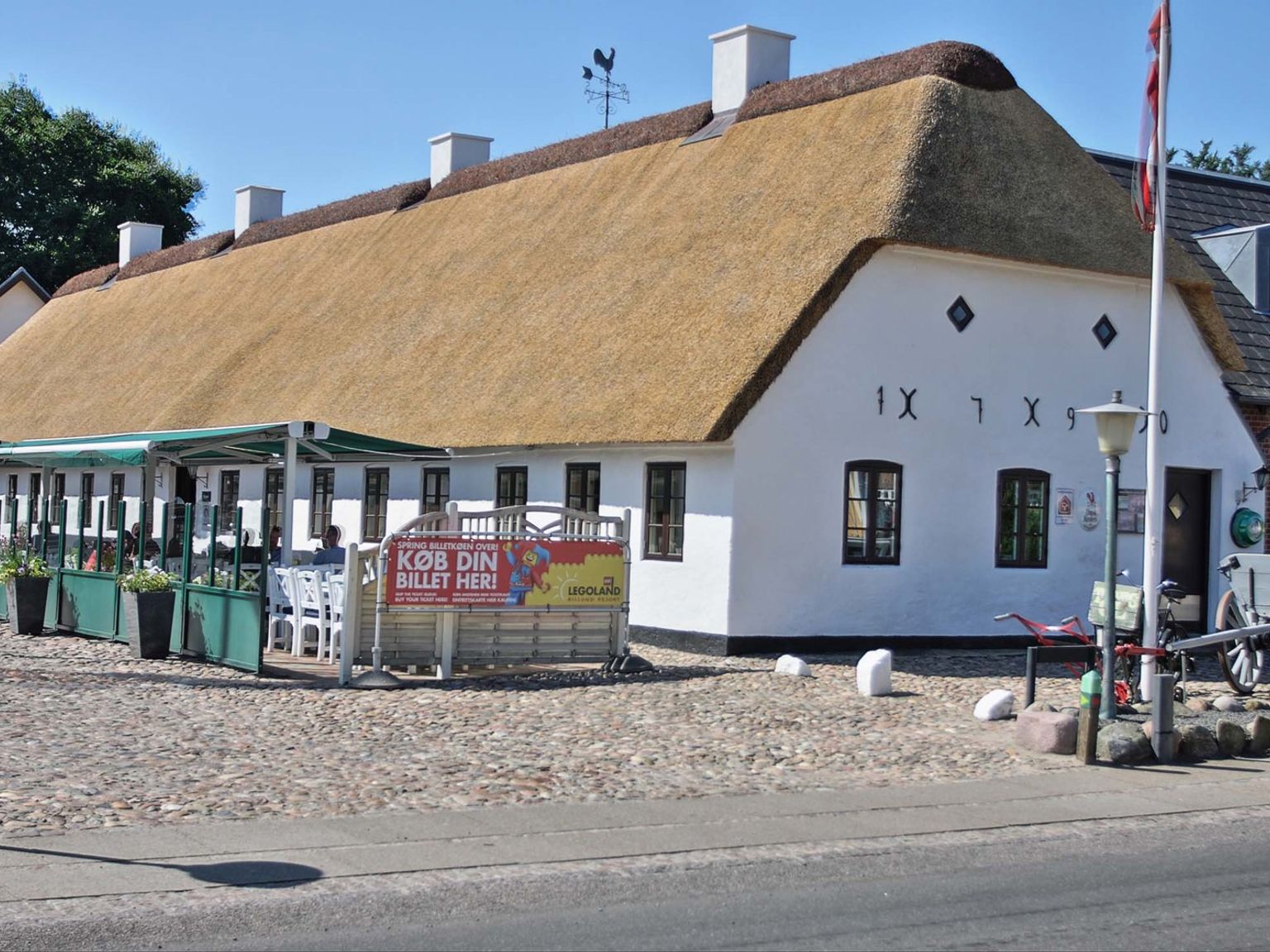 Ein langes, weißes Gebäude mit Reetdach und einem Außenbereich zum Essen auf einer Kopfsteinpflasterterrasse. Hovborg Kro