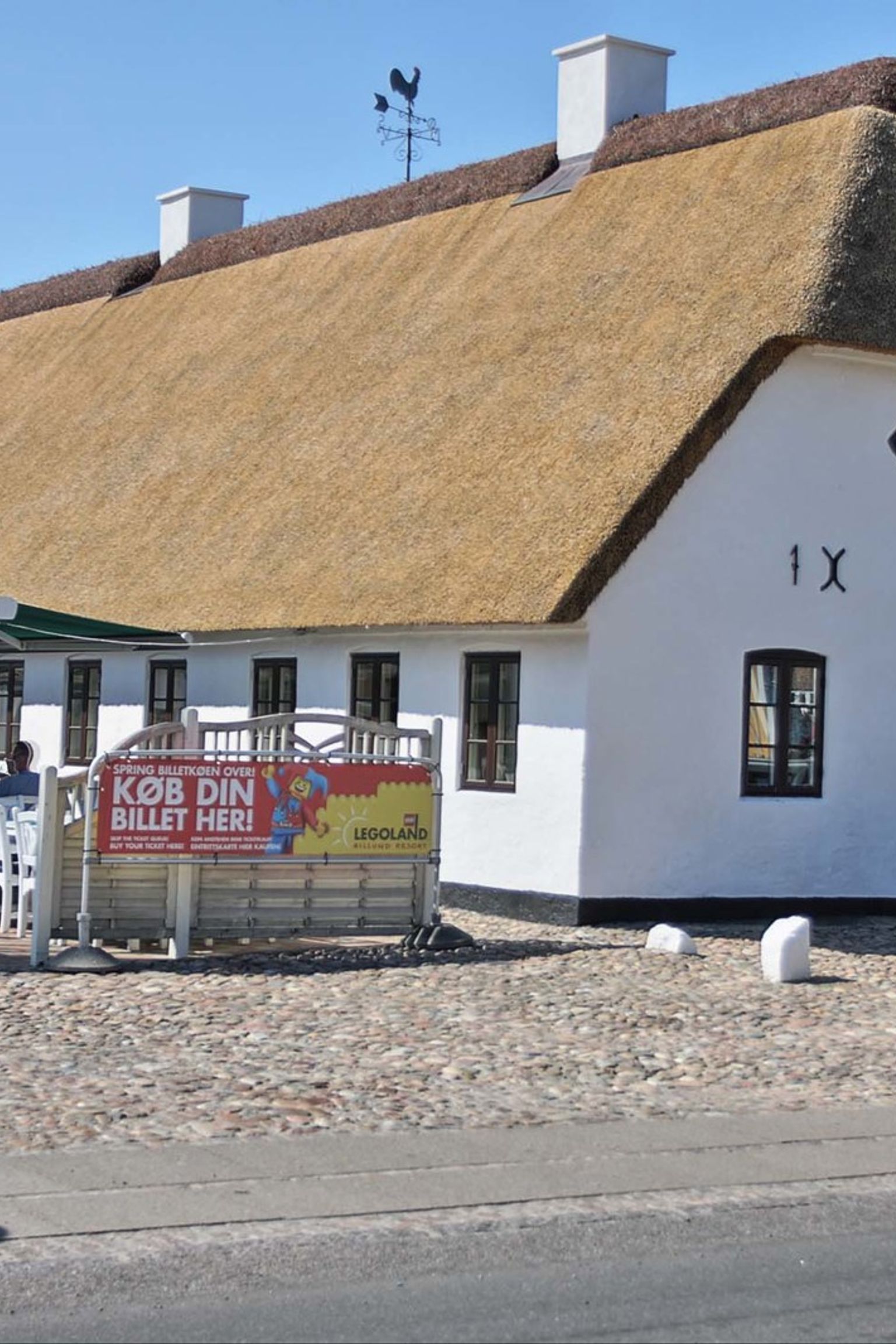 Ein langes, weißes Gebäude mit Reetdach und einem Außenbereich zum Essen auf einer Kopfsteinpflasterterrasse. Hovborg Kro