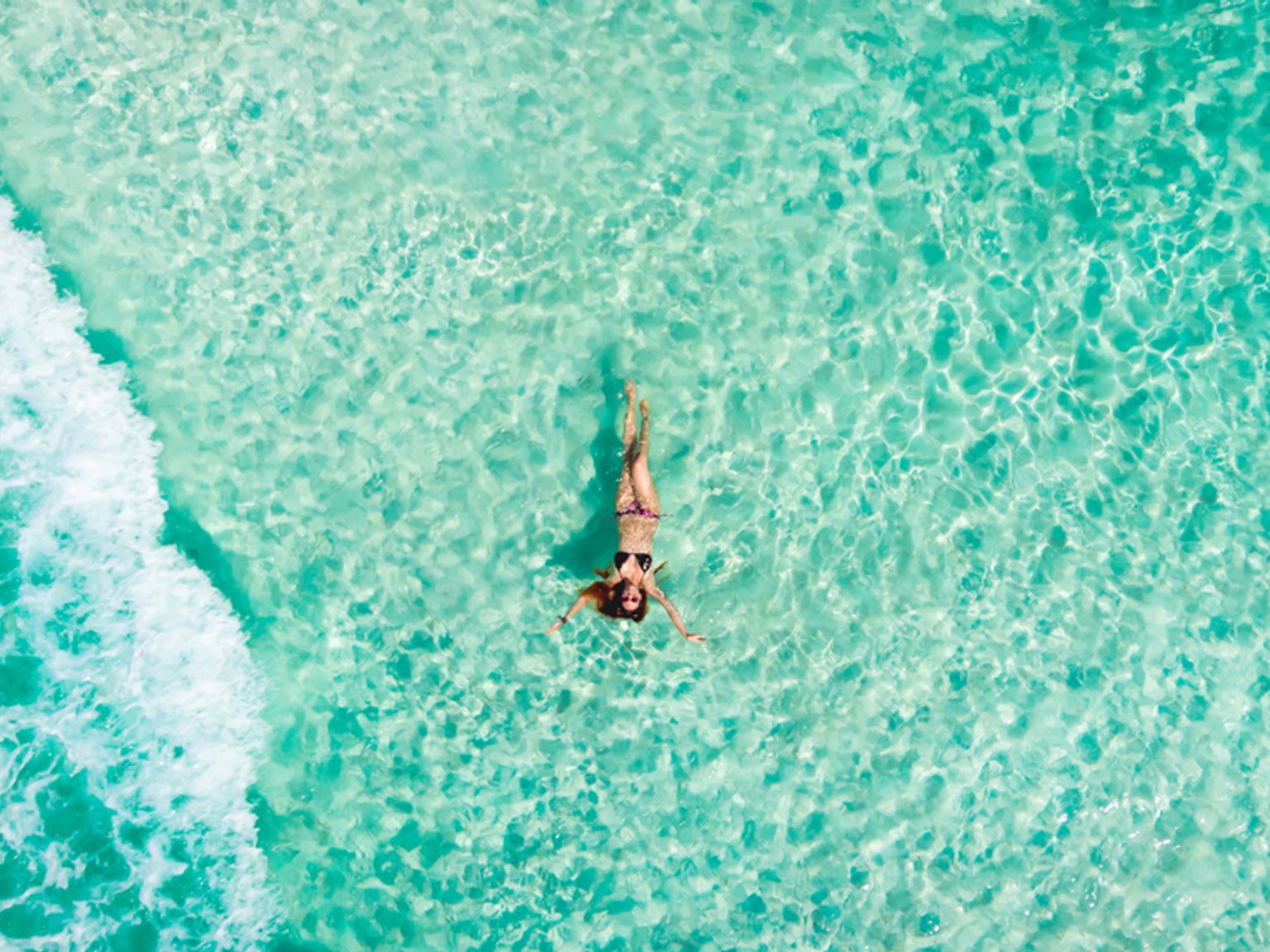 Flygfoto av en kvinna som flyter i klart turkost havsvatten bredvid en våg.