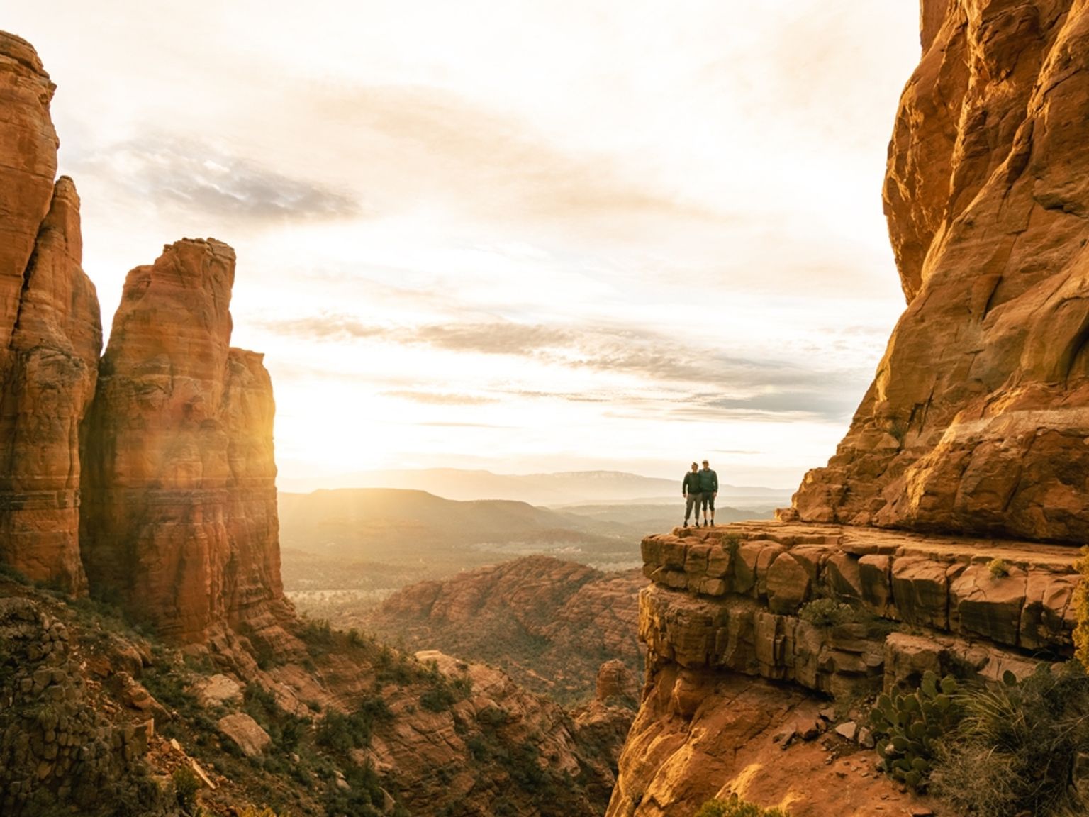 To personer står på en rød klippe og ser ut over en vidstrakt solbelyst canyon med tårnende formasjoner ved solnedgang. Sedona