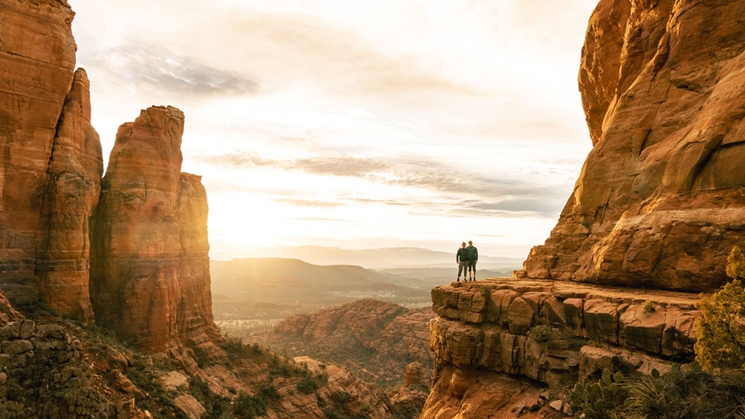 To personer stÄr pÄ en rÞd klippe og ser ut over en vidstrakt solbelyst canyon med tÄrnende formasjoner ved solnedgang. Sedona