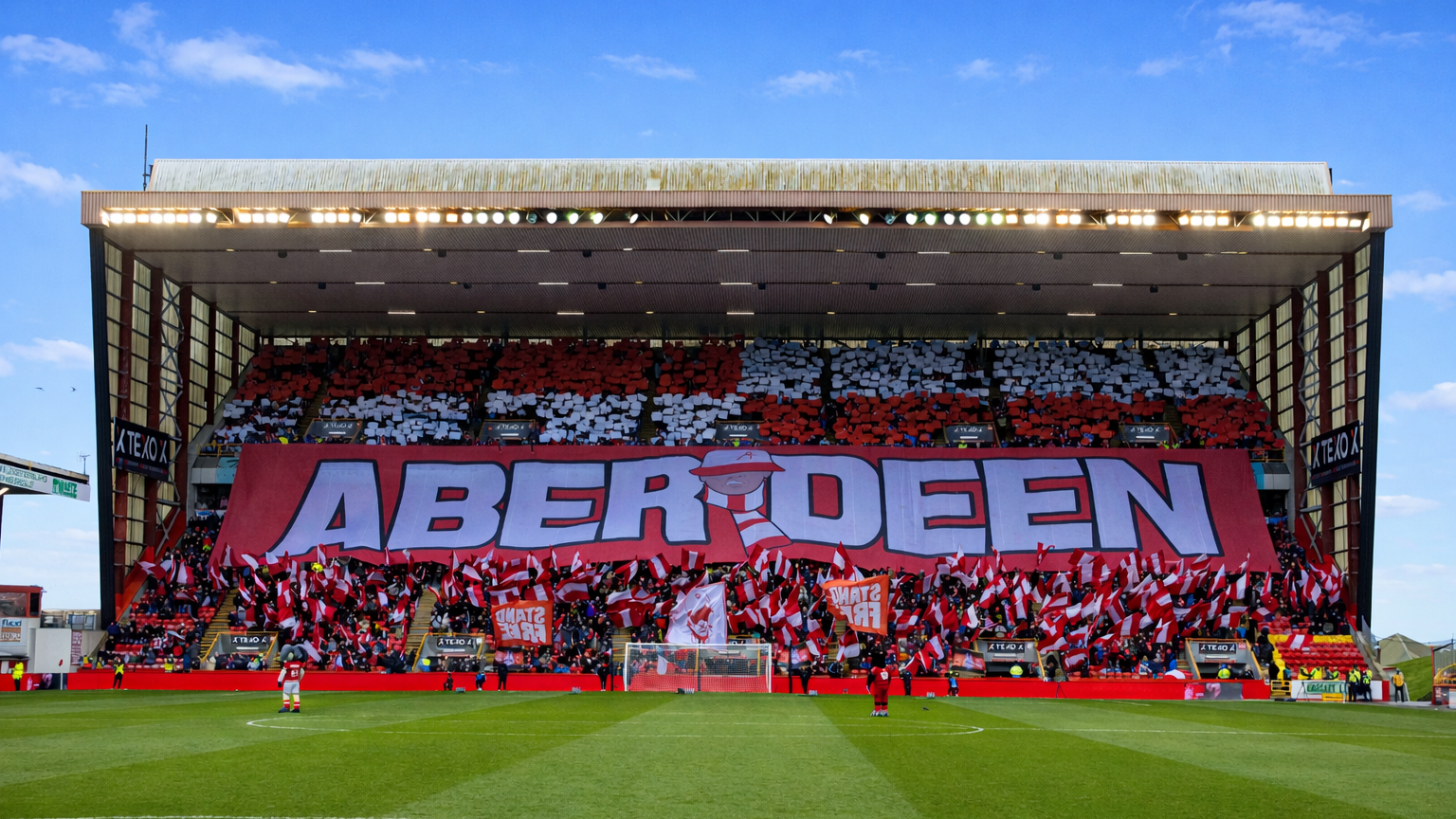 A packed football stadium stand features a massive "ABERDEEN" banner below rows of red and white fan card displays, with fans waving flags below.