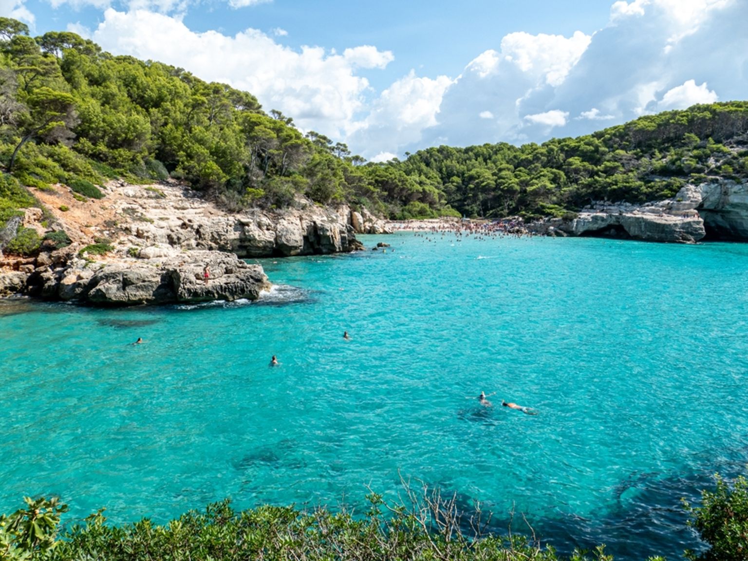 En livlig turkos vik med en trång strand och simmare, inramad av frodiga gröna klippor. Cala Mitjana, Menorca
