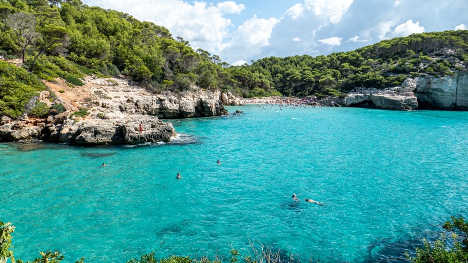 En livlig turkos vik med en trång strand och simmare, inramad av frodiga gröna klippor. Cala Mitjana, Menorca
