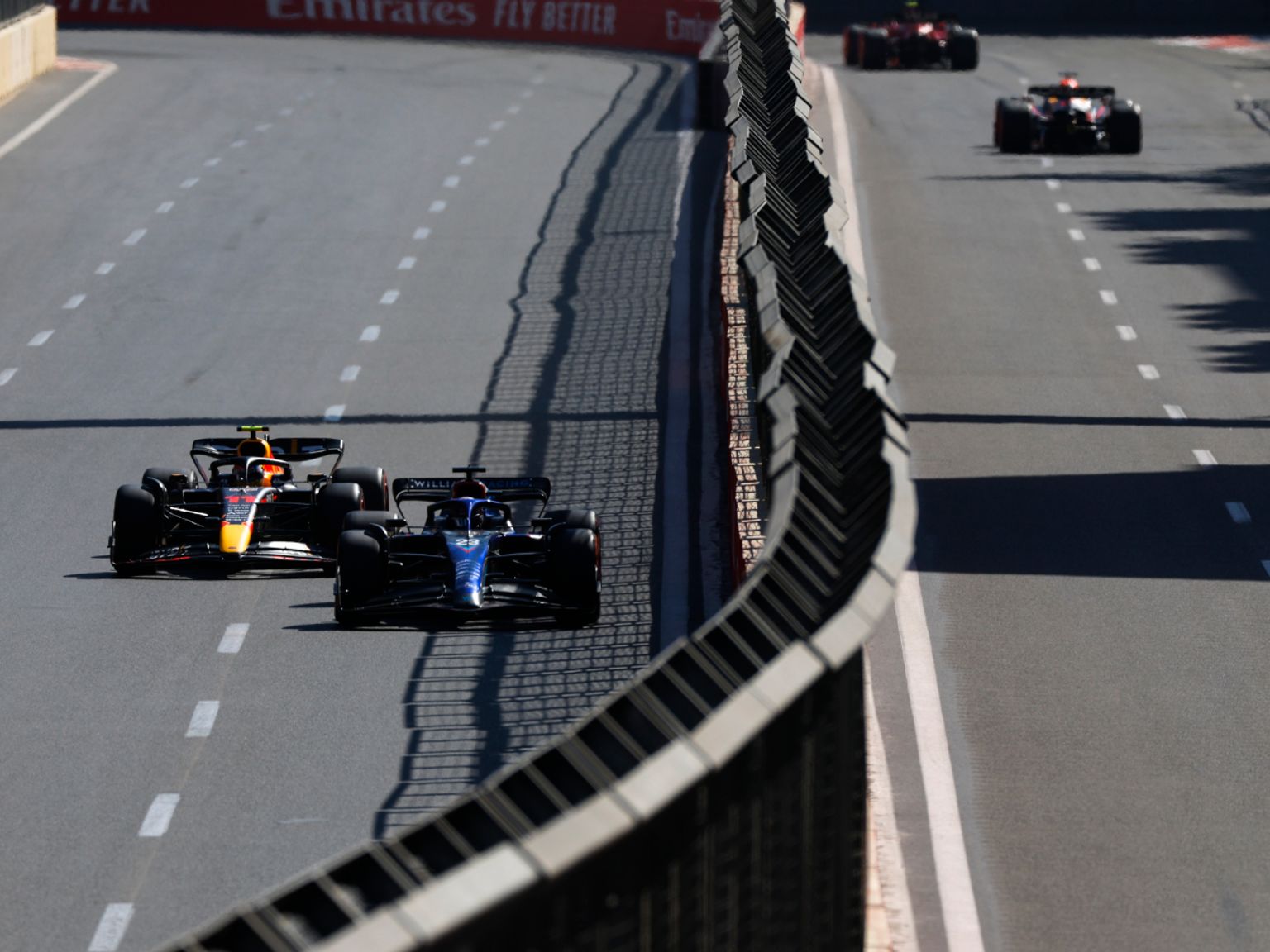 Sininen ja punainen-oranssi Formula 1 -auto kilpailevat lähekkäin aurinkoisella radalla, ja muita autoja näkyy edempänä.