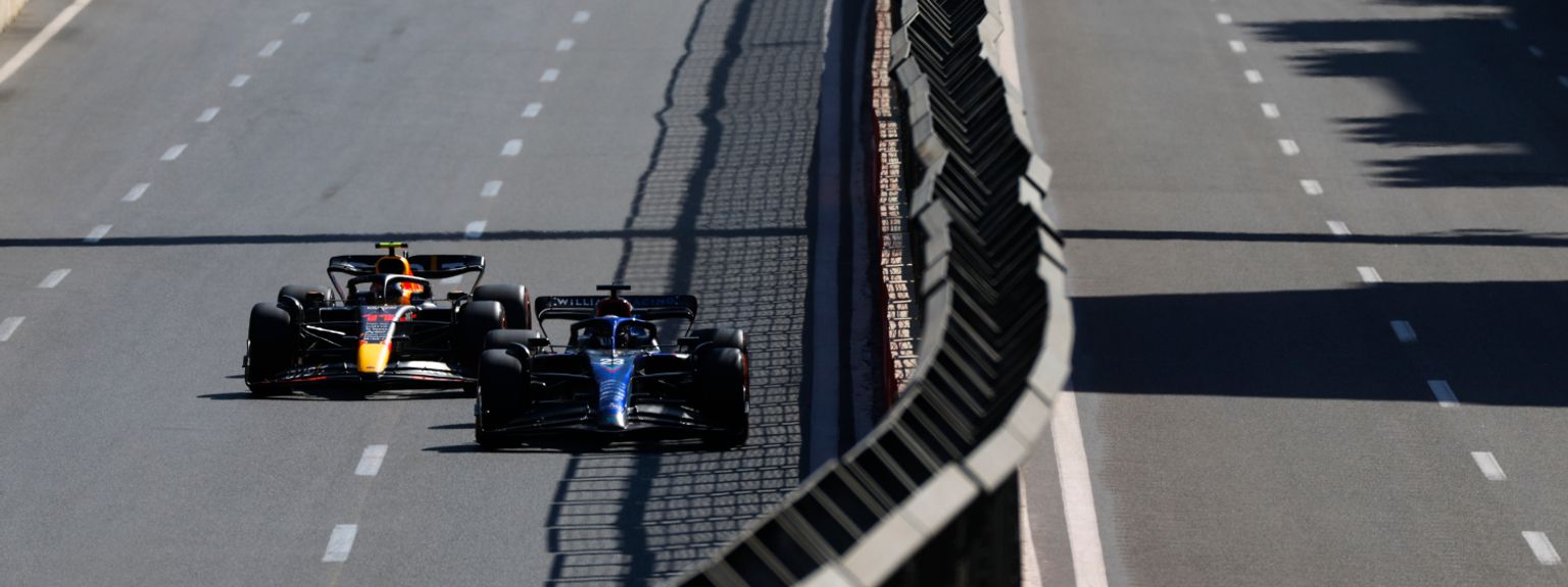 Sininen ja punainen-oranssi Formula 1 -auto kilpailevat lähekkäin aurinkoisella radalla, ja muita autoja näkyy edempänä.