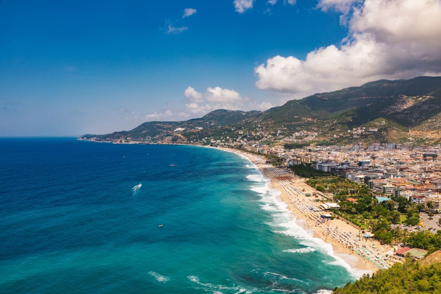 Luftfoto af en lang sandstrand med parasoller, en kystby og bjerge under en blå himmel. Alanya, Tyrkiet