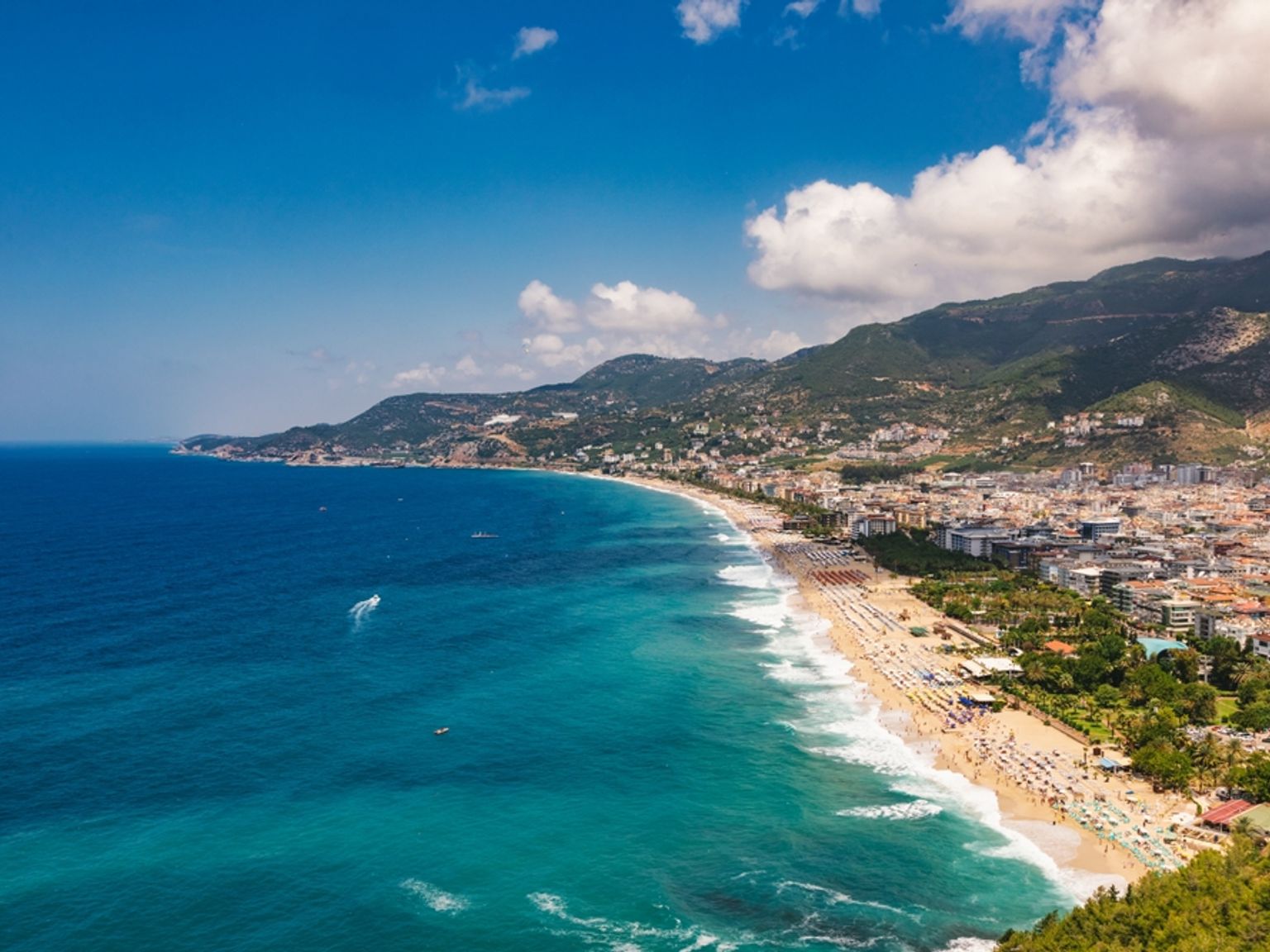 Luftfoto af en lang sandstrand med parasoller, en kystby og bjerge under en blå himmel. Alanya, Tyrkiet