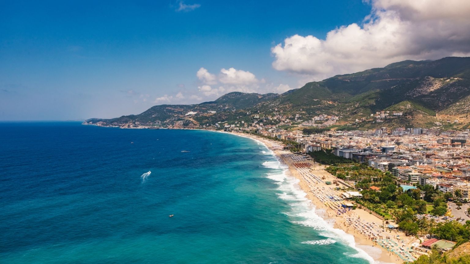 Luftfoto af en lang sandstrand med parasoller, en kystby og bjerge under en blå himmel. Alanya, Tyrkiet