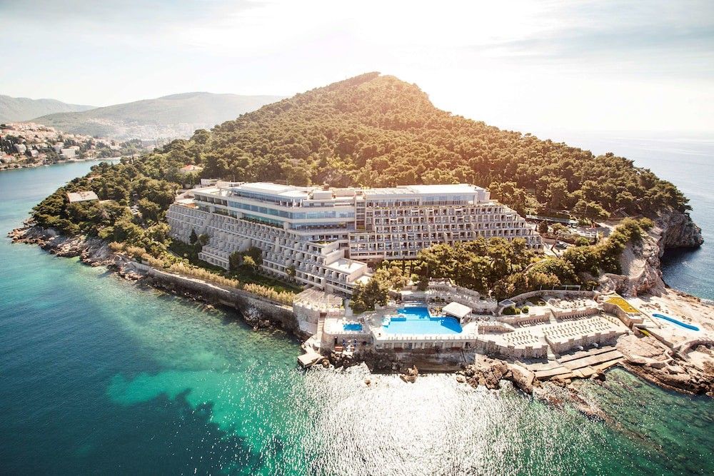Aerial view of a large terraced hotel complex on a wooded peninsula, surrounded by blue ocean with pools and sun decks. Dubrovnik Palace