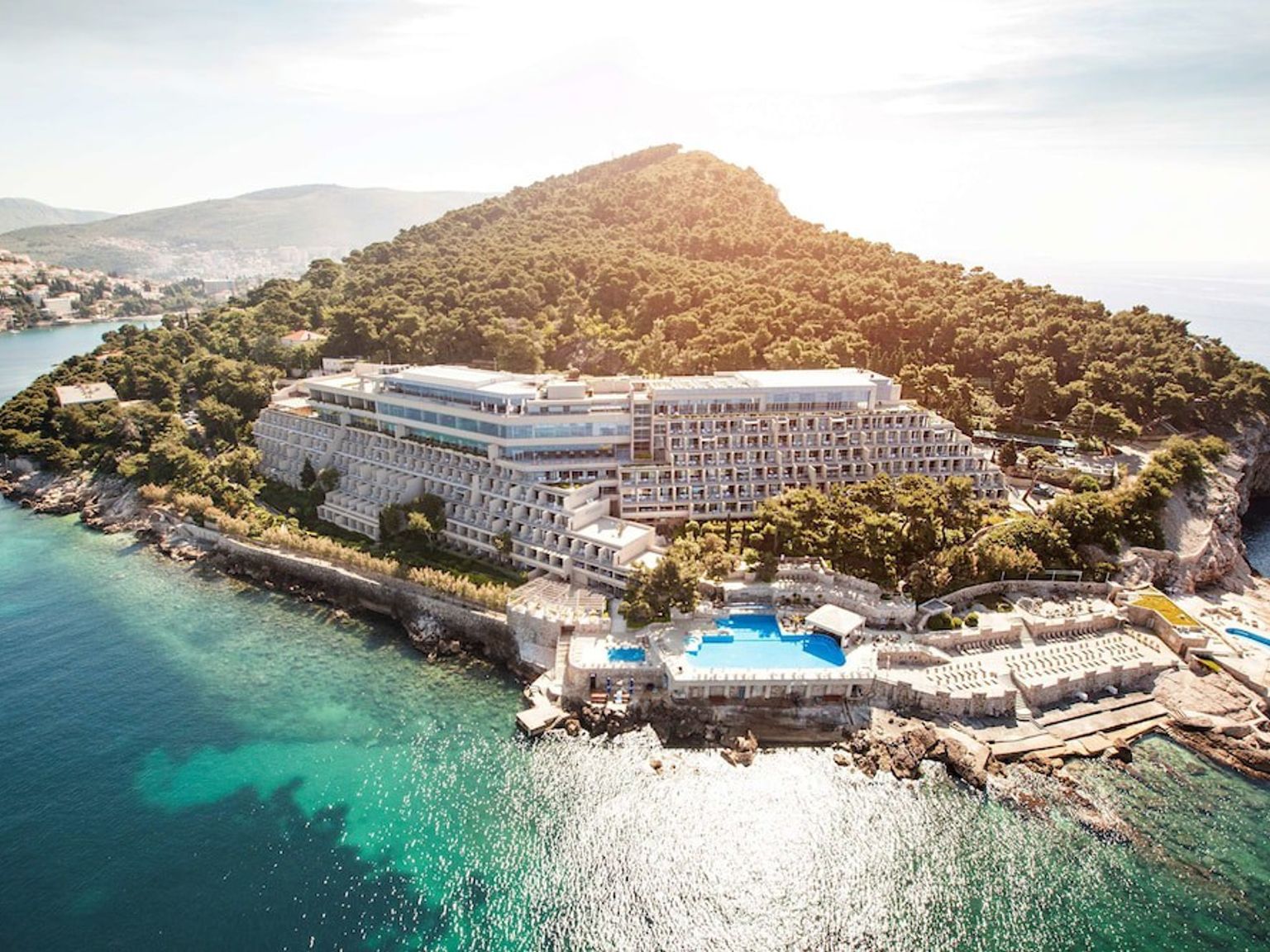 Aerial view of a large terraced hotel complex on a wooded peninsula, surrounded by blue ocean with pools and sun decks. Dubrovnik Palace