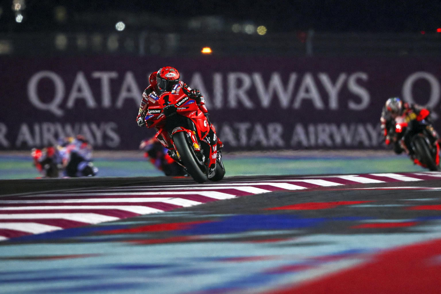 Un piloto de motocicleta rojo se inclina en una curva en una pista por la noche, con "QATAR AIRWAYS" visible al fondo.