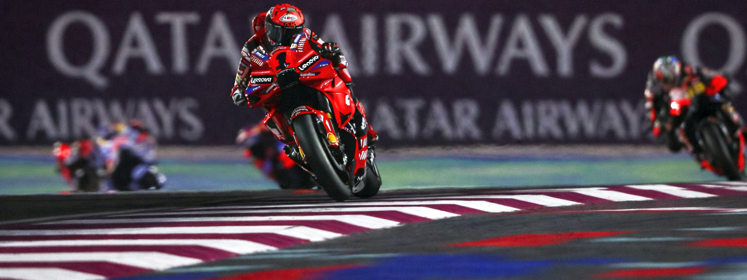 Un piloto de motocicleta rojo se inclina en una curva en una pista por la noche, con "QATAR AIRWAYS" visible al fondo.