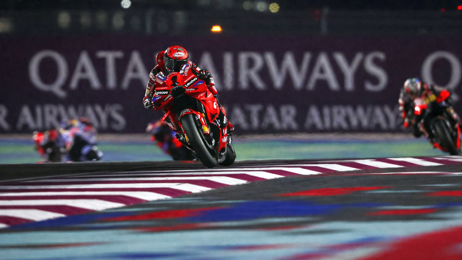 Un piloto de motocicleta rojo se inclina en una curva en una pista por la noche, con "QATAR AIRWAYS" visible al fondo.