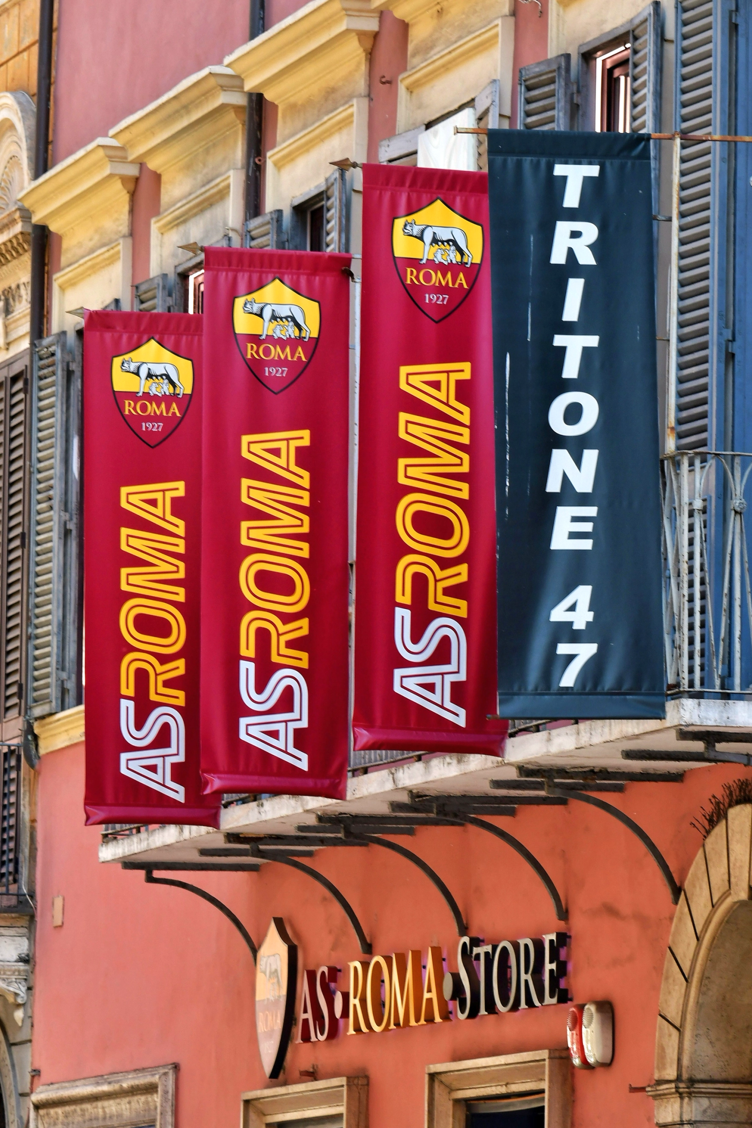 Røde AS Roma-bannere og et svart Tritone 47-banner henger fra en bygning over et AS Roma Store-skilt.