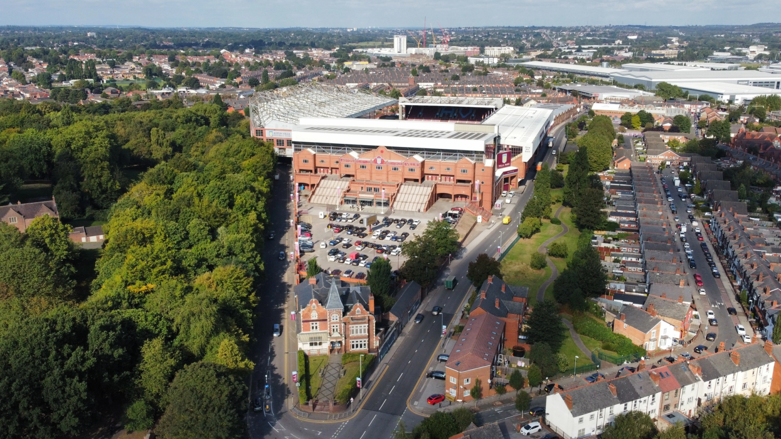 Aerial view of a large football stadium with brick-red walls and a white roof, bordered by dense green trees and residential streets with parked cars.