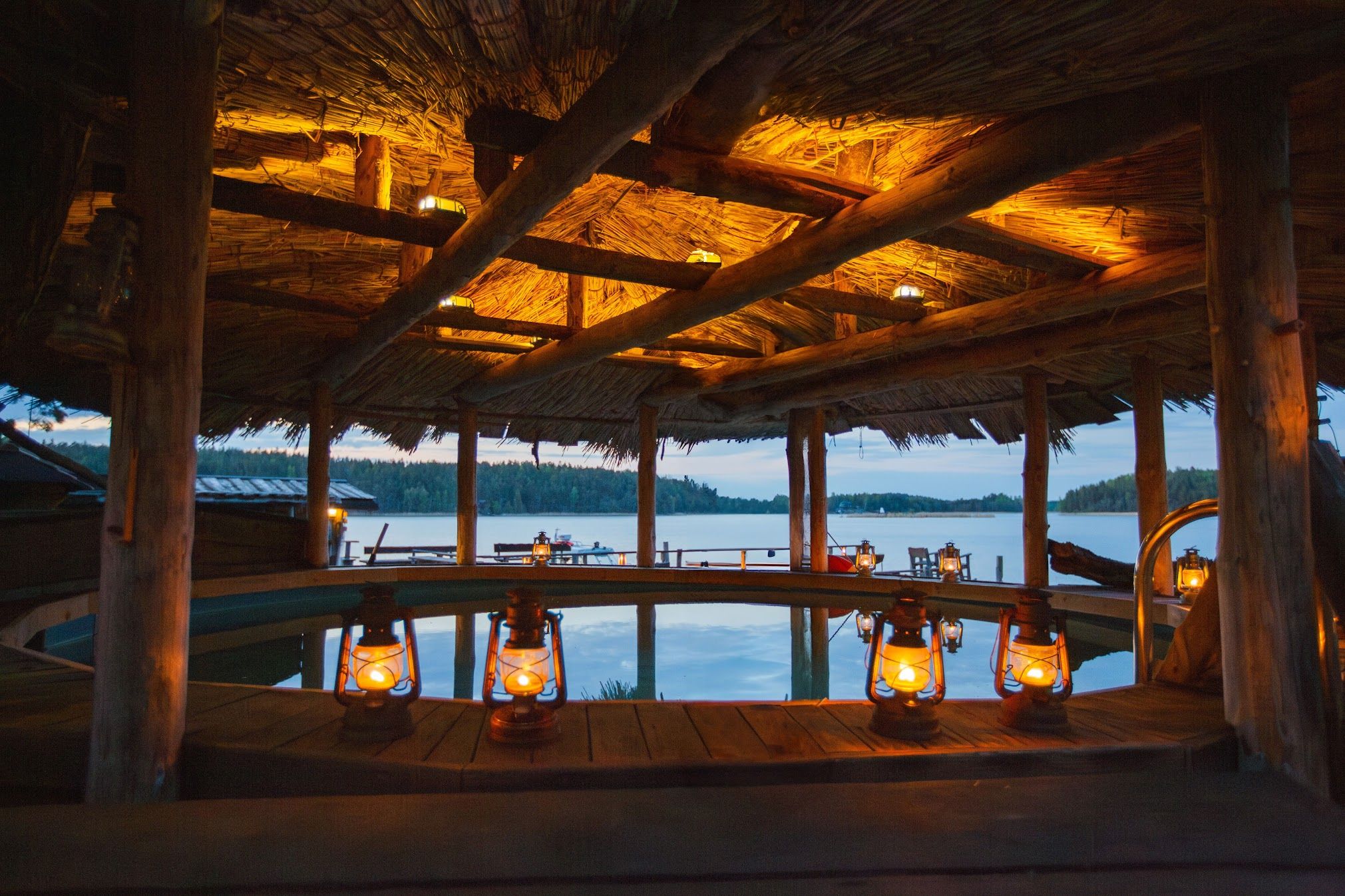 Et rustikt udendørs vildmarksbad med tændte lanterner under et stråtag med udsigt over en sø i skumringen.