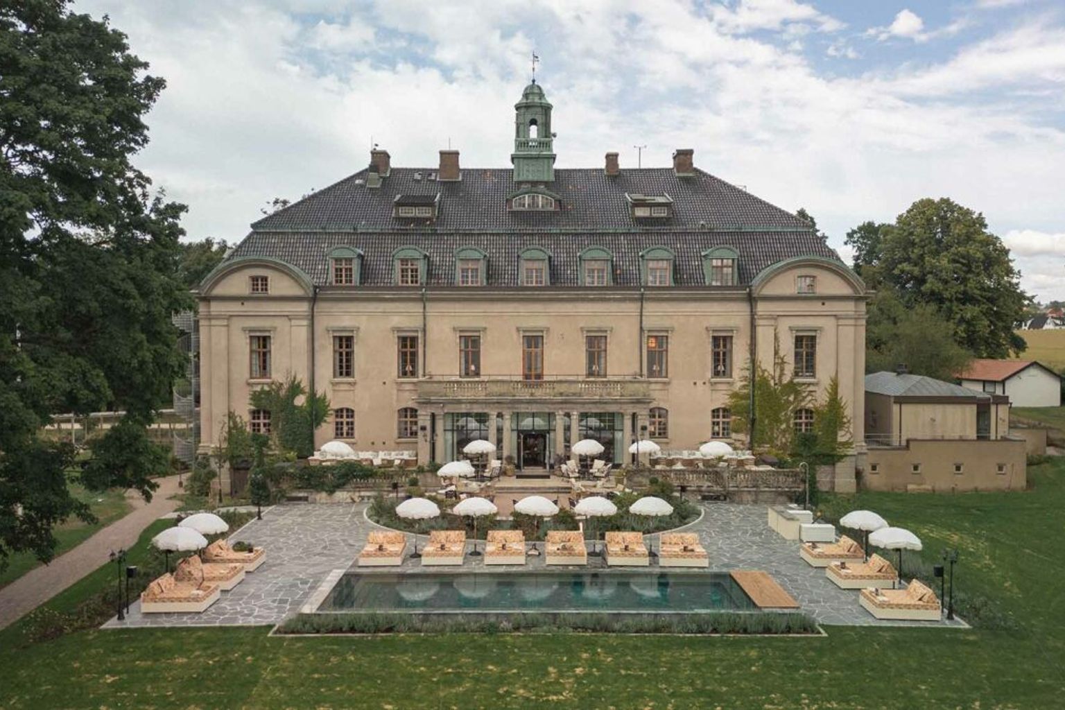 En storslagen byggnad med en utomhuspool, uteplats, solstolar och vita parasoller på en grön gräsmatta. Maryhill Estate