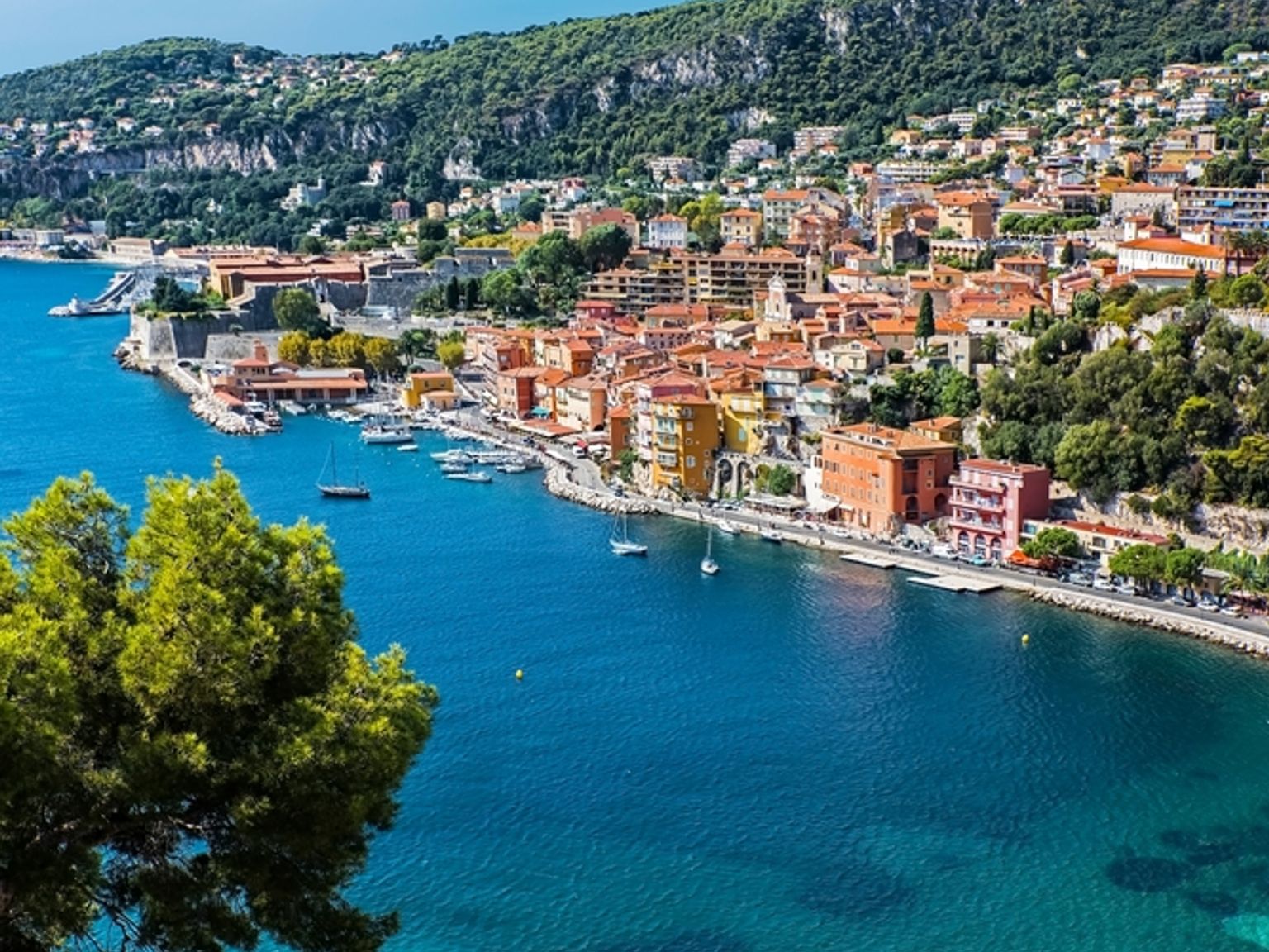 Eloisa rannikkokylä, jossa on värikkäitä rakennuksia turkoosin lahden varrella, hiekkaranta, vihreitä kukkuloita ja vaaleanpunaista bougainvilleaa etualalla. Villefranche-sur-Mer, Ranska