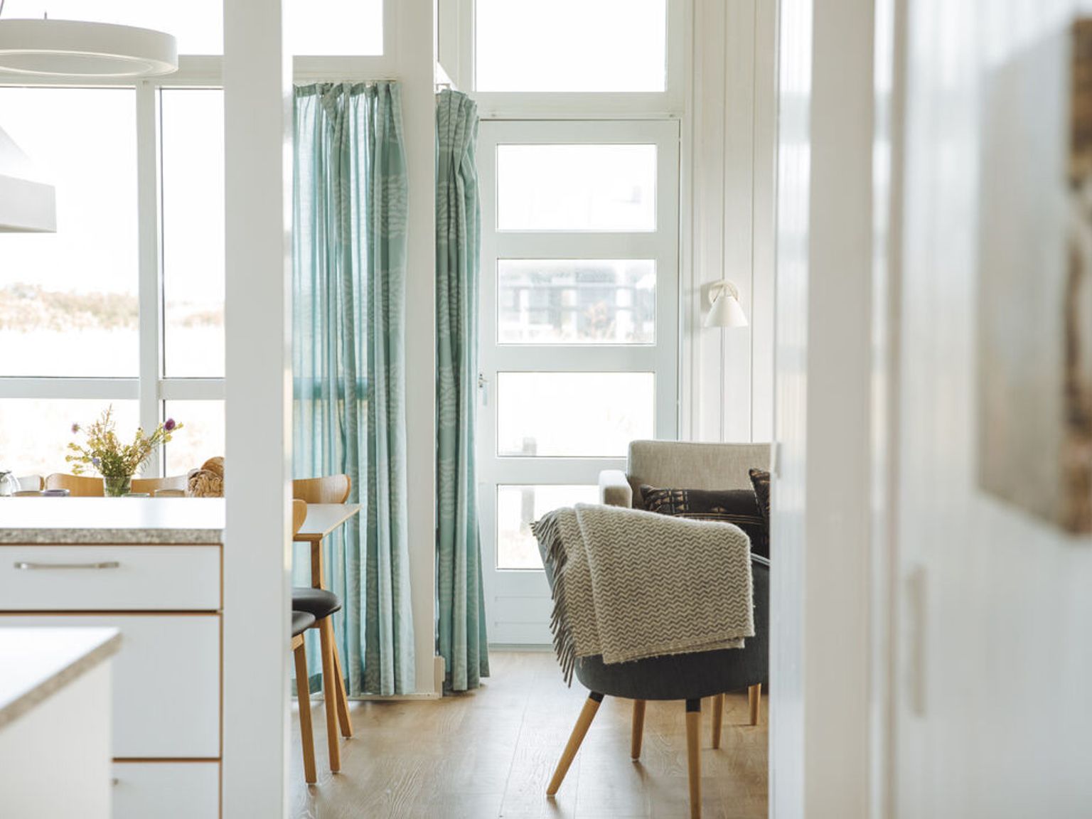 Lyst hjem interiør med kjøkkenbenk, spisebord og lenestol i stuen, med store vinduer og blå gardiner.