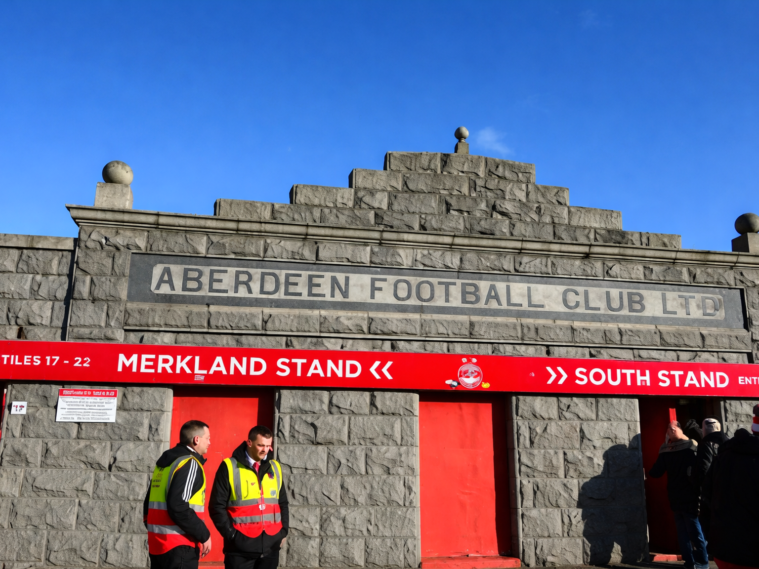 Stenfacadeindgang til Aberdeen Football Club stadion med dets navn indgraveret over røde tribuneskilte, og folk samlet nedenfor.