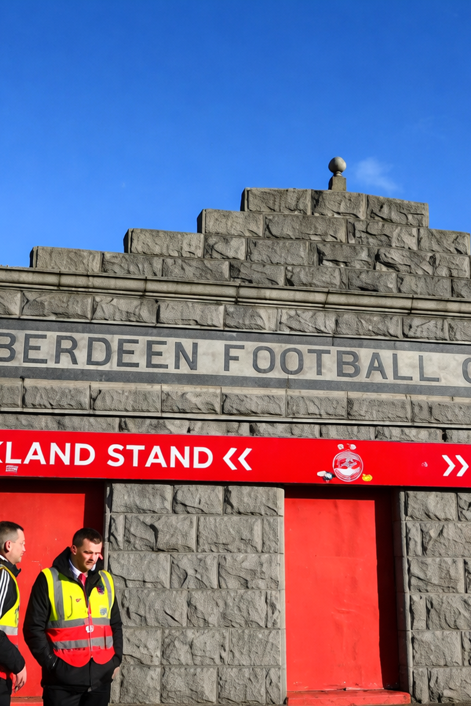 Stenfacadeindgang til Aberdeen Football Club stadion med dets navn indgraveret over røde tribuneskilte, og folk samlet nedenfor.