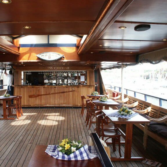 Træloungebåd med baren "Princess Caroline", små borde og dækpladser med udsigt over en marina.