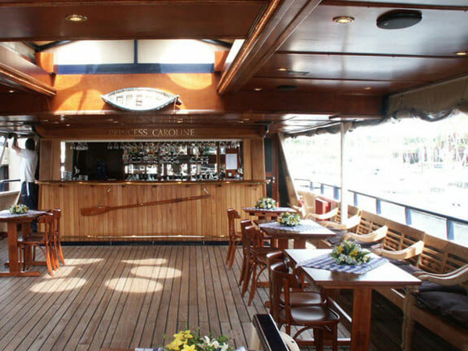 Træloungebåd med baren "Princess Caroline", små borde og dækpladser med udsigt over en marina.