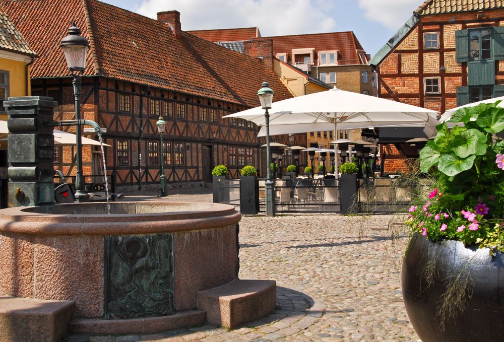 Ett kullerstensbelagt torg med en fontän, korsvirkeshus och ett utomhuscafé.