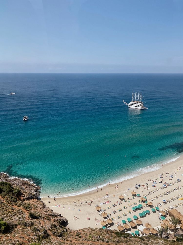 En højvinkeludsigt over en solrig strand med turkis vand, mange strandgæster, parasoller og en stor hvid sejlbåd i havet. Alanya, Tyrkiet