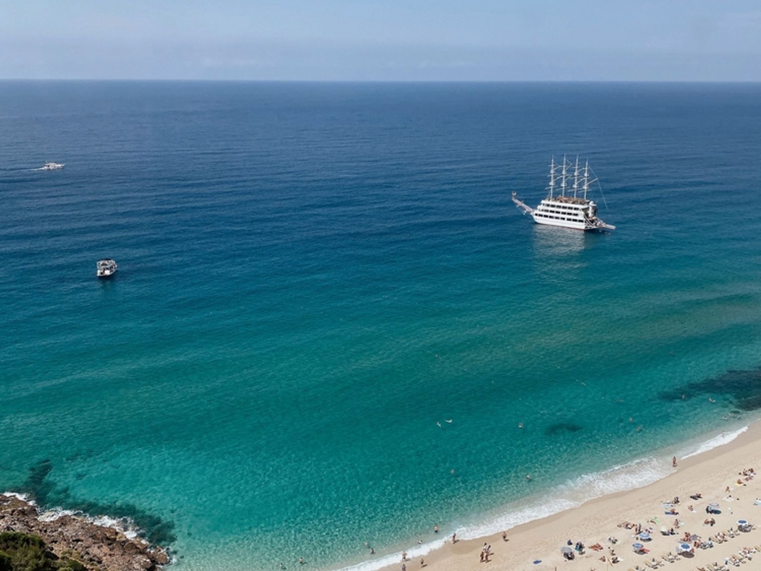 En højvinkeludsigt over en solrig strand med turkis vand, mange strandgæster, parasoller og en stor hvid sejlbåd i havet. Alanya, Tyrkiet