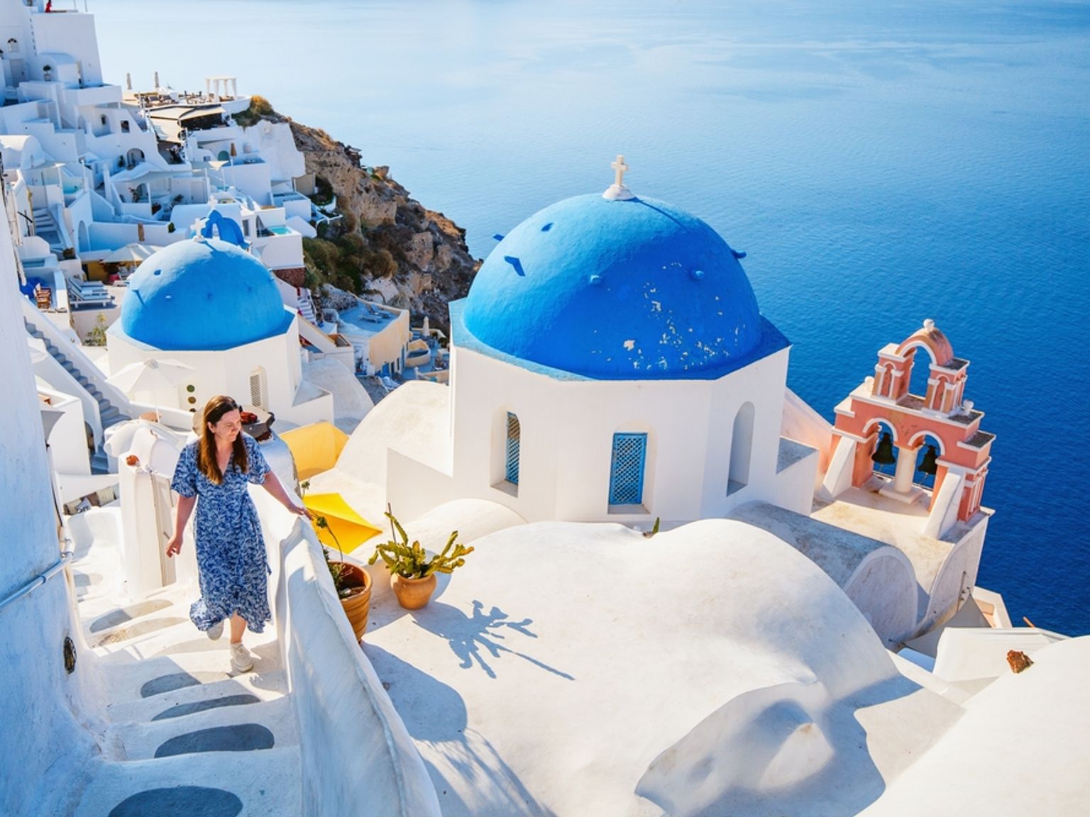 En kvinna går nerför vita trappor bland blåkupolerade kyrkor och vita byggnader med utsikt över det blå havet i Santorini.
