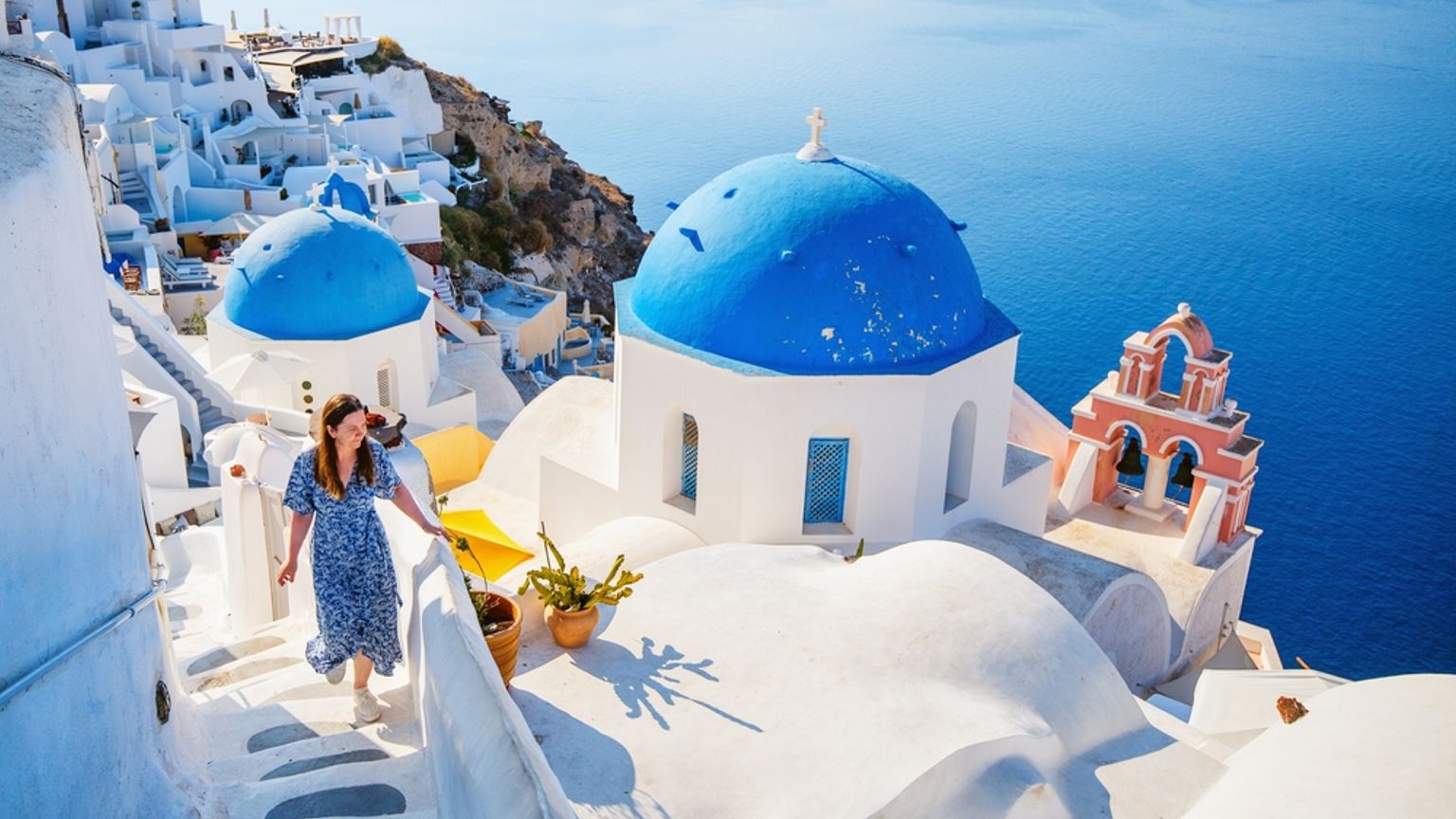 En kvinde går ned ad hvide trapper blandt blåkupolerede kirker og hvide bygninger med udsigt over det blå hav i Santorini.
