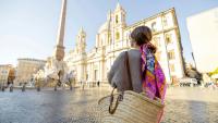 Rückansicht einer Frau mit einem bunten Schal und Strohtasche, die die Piazza Navona in Rom bewundert, mit einer Barockkirche, einem Obelisken und einem Brunnen.