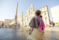 Rückansicht einer Frau mit einem bunten Schal und Strohtasche, die die Piazza Navona in Rom bewundert, mit einer Barockkirche, einem Obelisken und einem Brunnen.