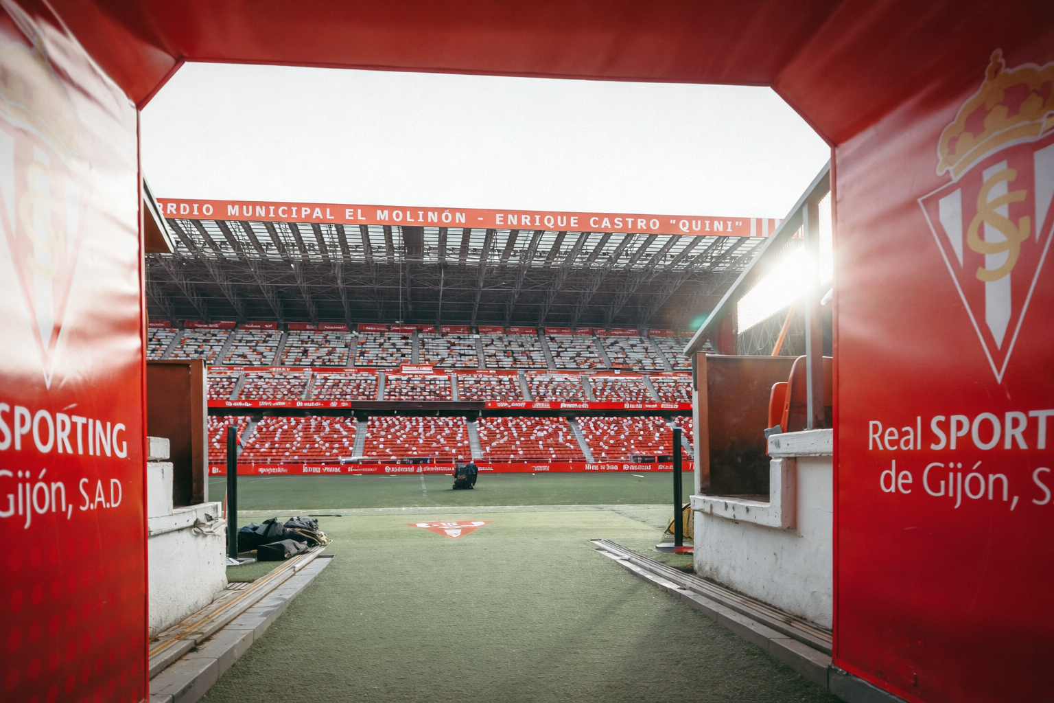 View from a red stadium tunnel onto the football pitch and the empty red seats of Estadio El Molinón, branded with Sporting Gijón.