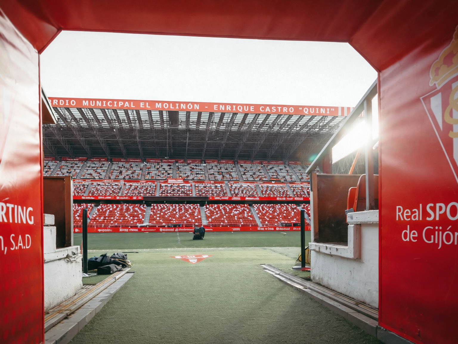 View from a red stadium tunnel onto the football pitch and the empty red seats of Estadio El Molinón, branded with Sporting Gijón.
