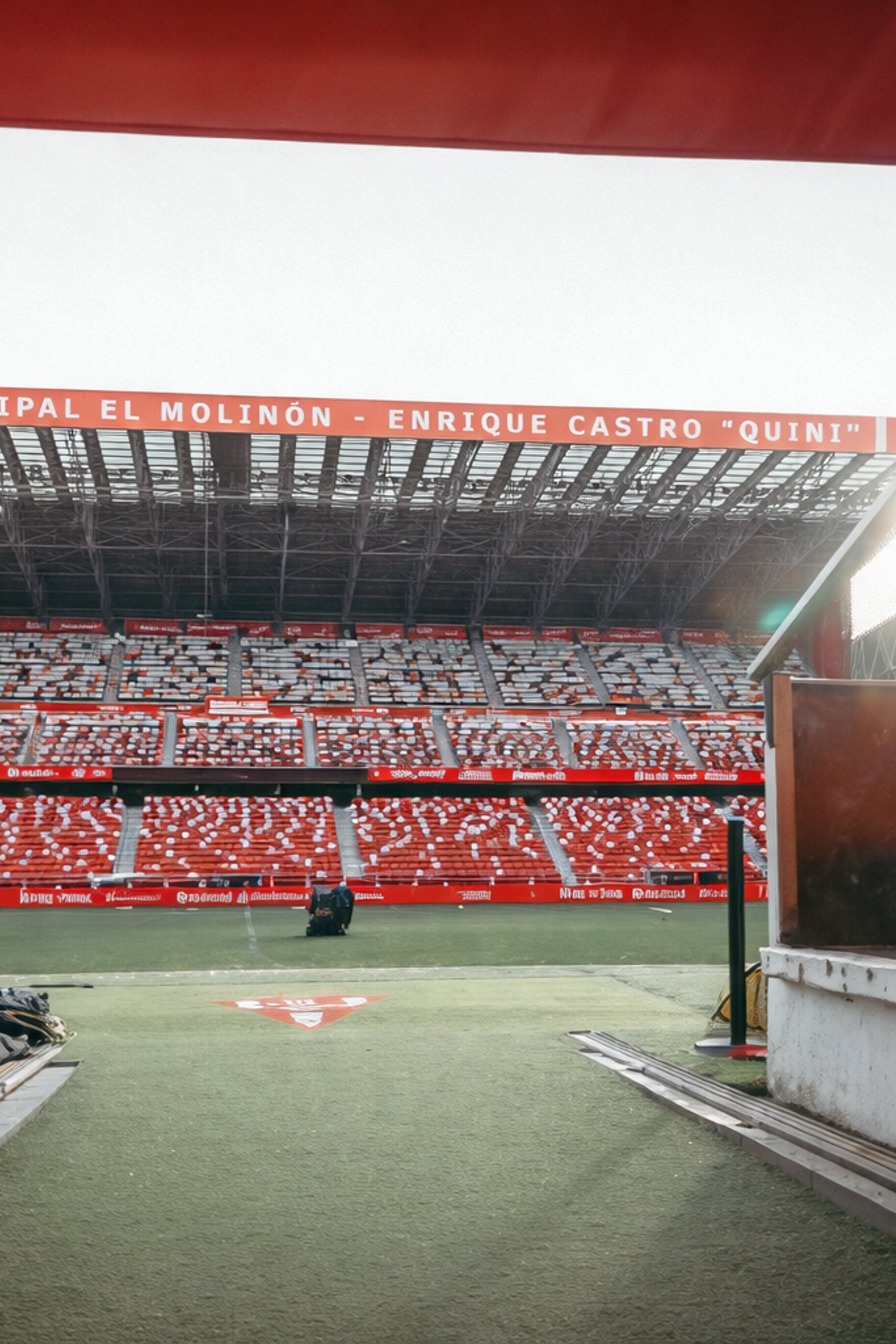 View from a red stadium tunnel onto the football pitch and the empty red seats of Estadio El Molinón, branded with Sporting Gijón.